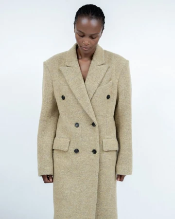 Fashionable woman in a tan double-breasted overcoat against a white background in a studio setting, looking down in a medium shot.