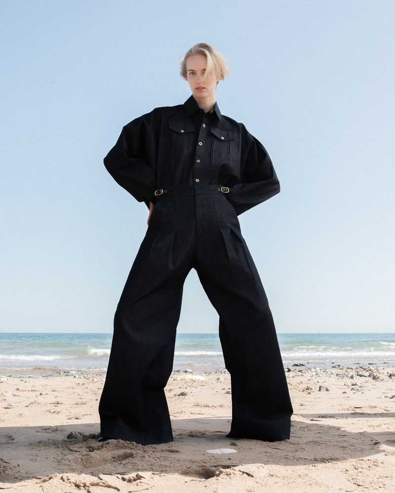 A fair-skinned woman poses on a beach in a dark denim jumpsuit, set against a light blue sky and ocean backdrop.