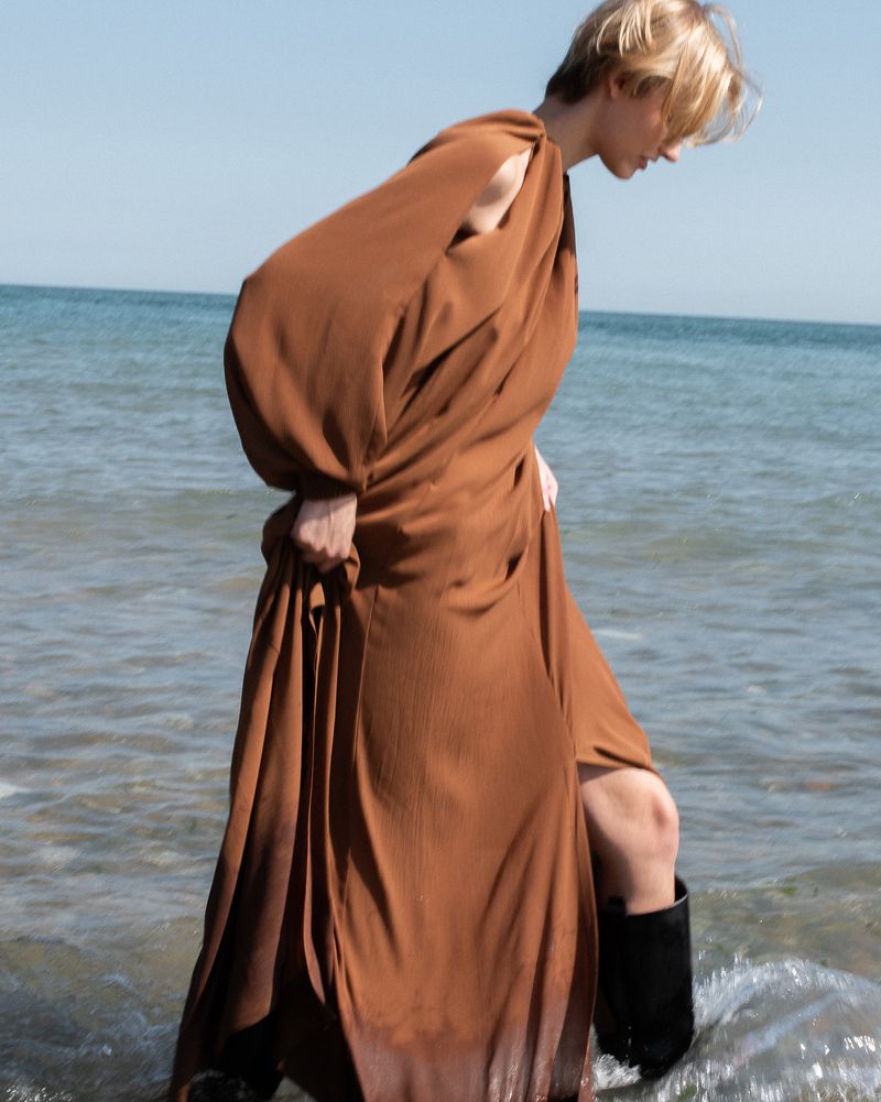 A woman stands in shallow ocean water wearing a brown dress and black boots on a sunny day, looking down with the blue ocean in the background.