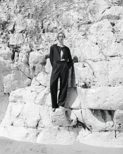 Model wearing a black cropped jacket and wide-leg trousers standing barefoot on a textured white rock cliff in black and white.