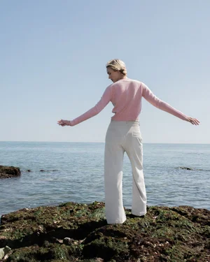 Model standing on a rocky shore wearing a pink sweater and pale trousers against the ocean
