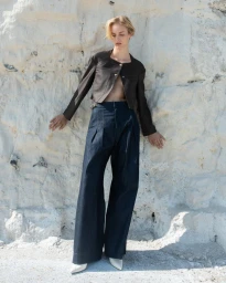 Model wearing a cropped brown jacket and pleated wide-leg dark denim trousers standing against a white rocky cliff wall.