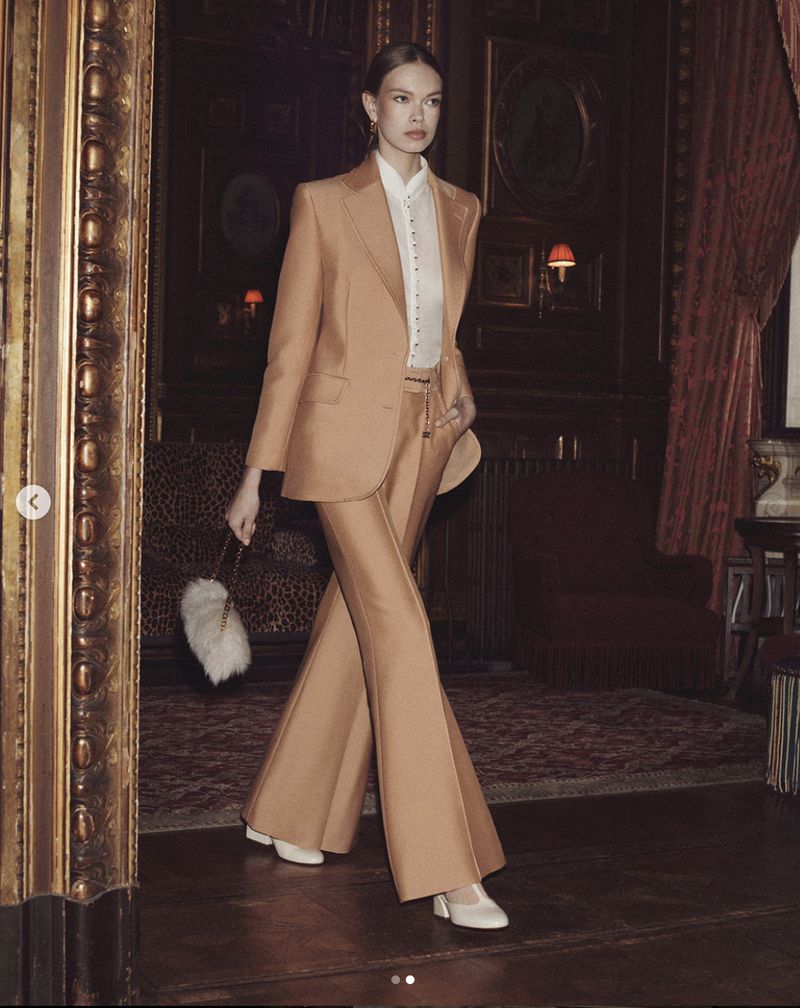 Adult female in a tan suit walks toward the camera inside an ornately decorated interior with dark wood and ornate details, holding a fuzzy handbag.