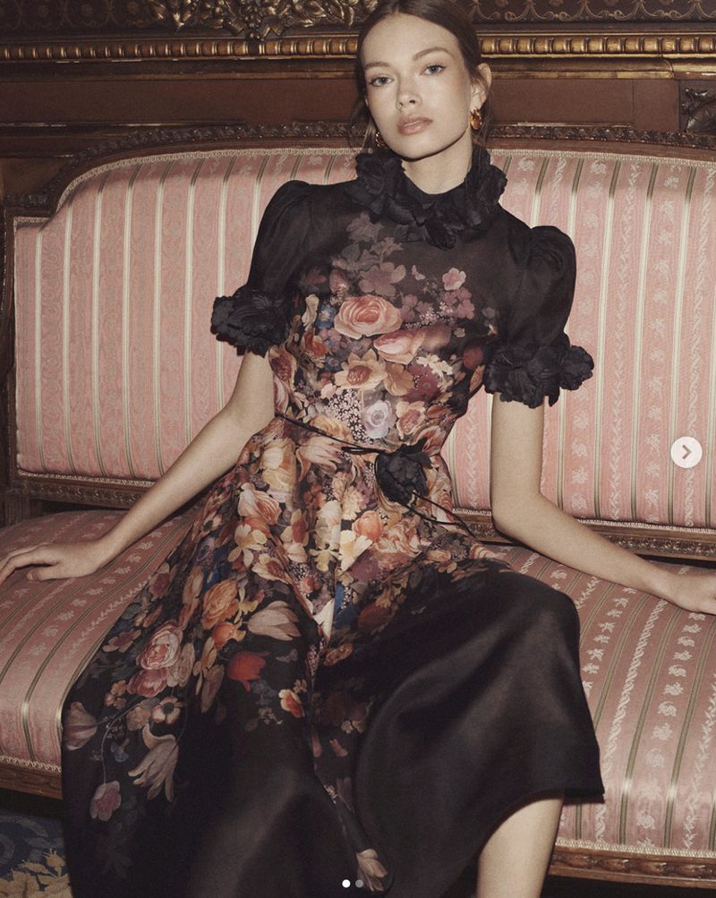 Adult woman wearing floral-print formal dress sitting on a pink and gold striped couch in a classically decorated interior.
