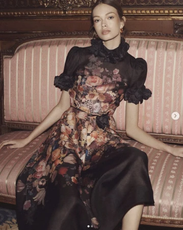 Adult woman wearing floral-print formal dress sitting on a pink and gold striped couch in a classically decorated interior.