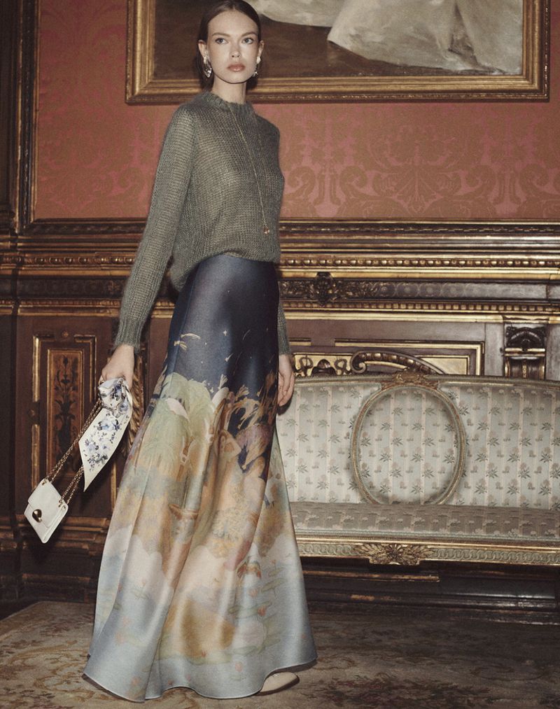 Model Sophie Bozhenova poses in a grey knit sweater and painterly printed maxi skirt inside an ornate room for Zimmermann FW23.