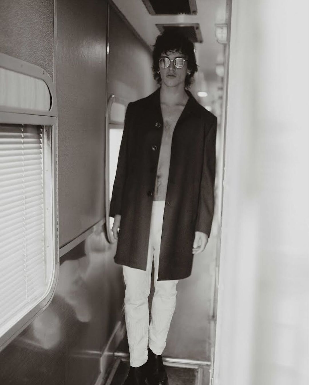 Monochrome portrait of a young adult male in a jacket and light pants standing in a train car with natural light and shadows.