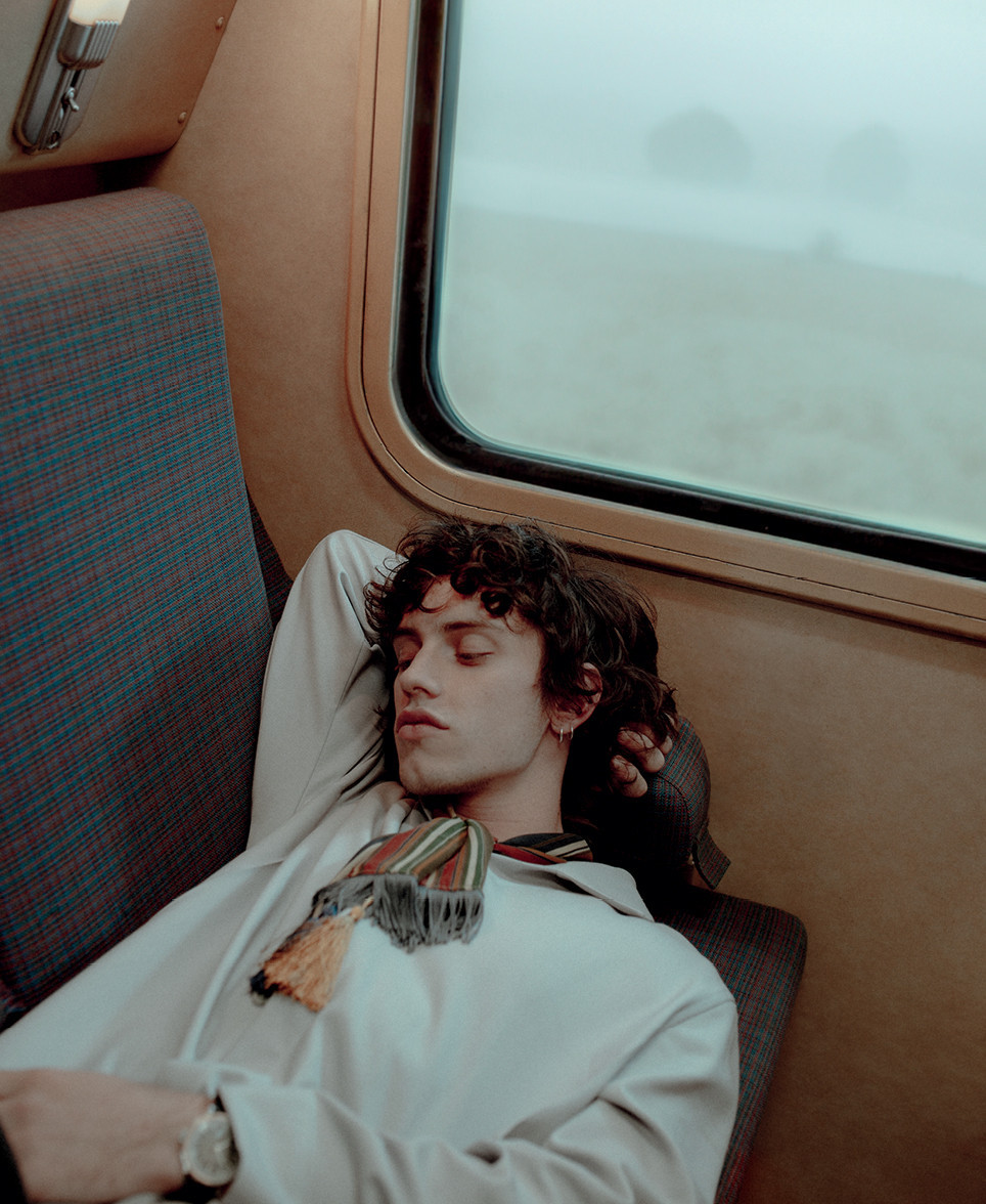Man asleep on train seat, wearing a light jacket and patterned scarf. The window shows a misty, blurred landscape outside.
