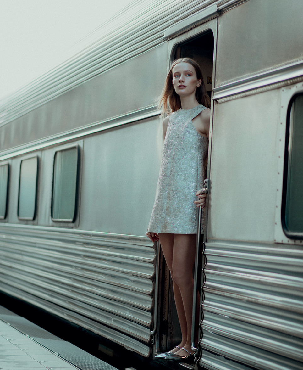A slender woman in a silver dress stands in a train doorway, gazing forward. The train's metallic exterior features horizontal lines.