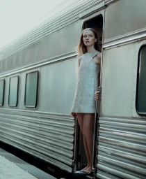A female model standing in the doorway of a vintage silver train carriage wearing a patterned dress.