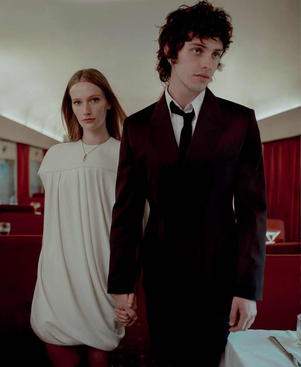 A stylish couple in a retro setting. The man wears a black suit, while the woman is in an elegant white dress, holding hands.