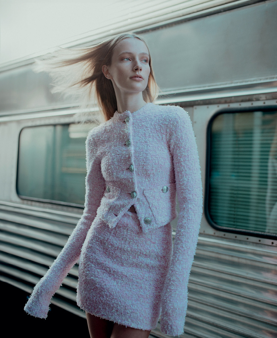 A slender woman in a pink tweed suit poses at a train station with a blurred metal train in the background and soft, diffused sunlight.