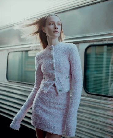 A slender woman in a pink tweed suit poses at a train station with a blurred metal train in the background and soft, diffused sunlight.