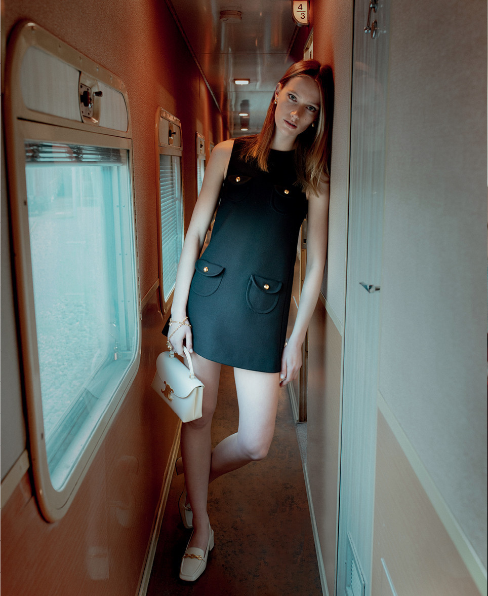 An adult female model poses in a short black dress with a white handbag in the narrow corridor of a passenger train interior.