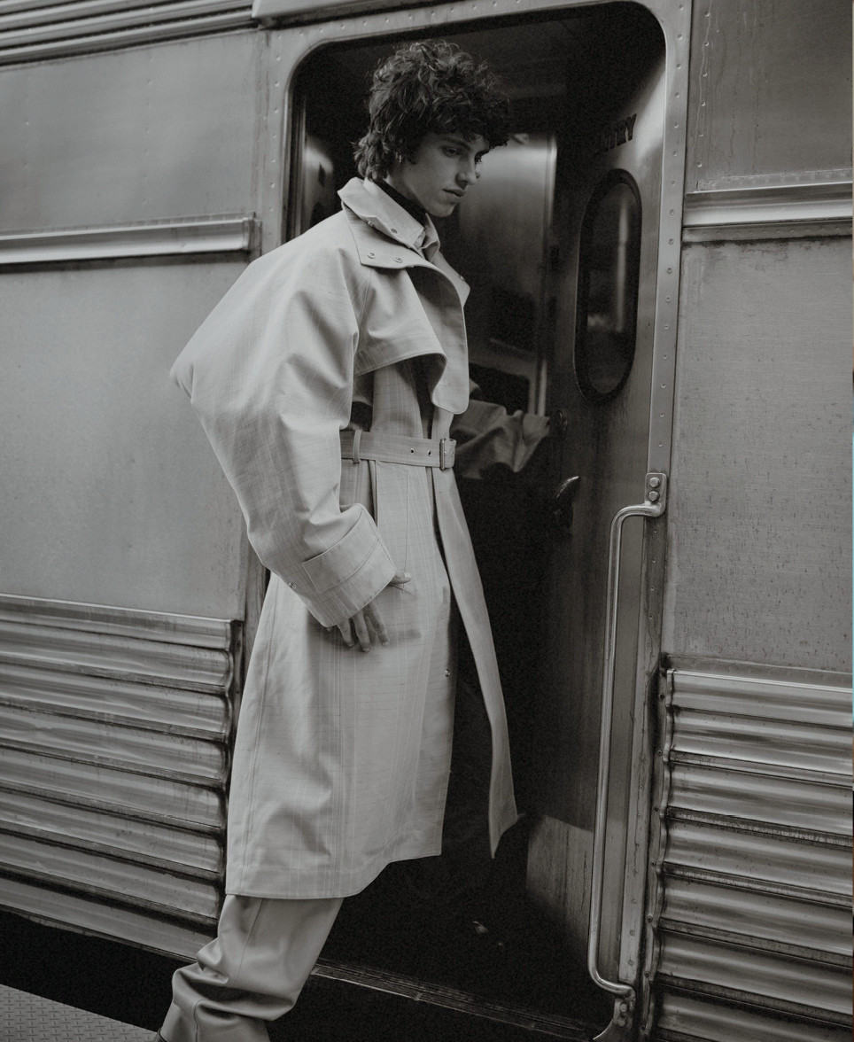 Monochrome image of a curly-haired adult male model in a trench coat exiting a vintage train car, showcasing a blend of fashion and travel.