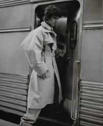 Young man in an oversized belted trench coat stepping out of a train carriage doorway in monochrome.