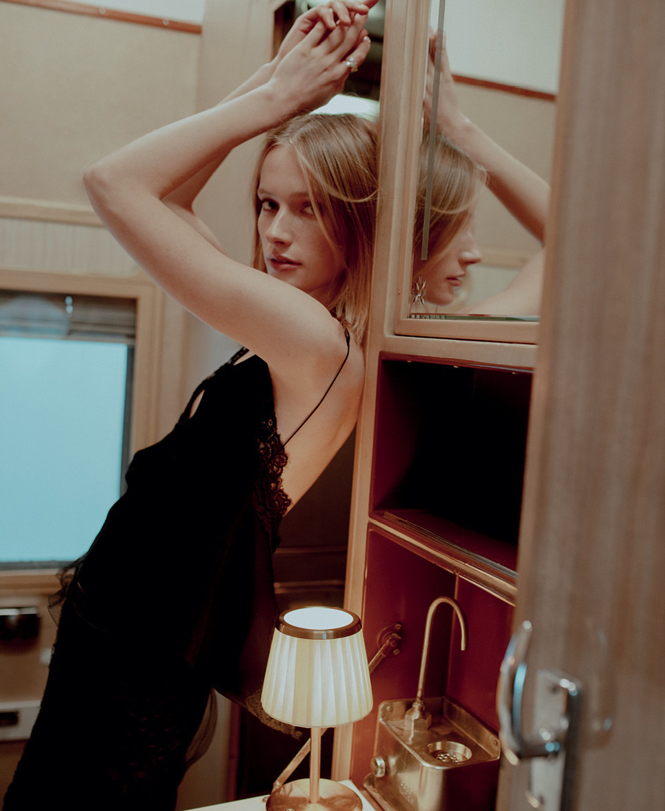 Woman in black dress leans against a wall with a mirror, showcasing fashion and beauty in an indoor setting with warm lighting.