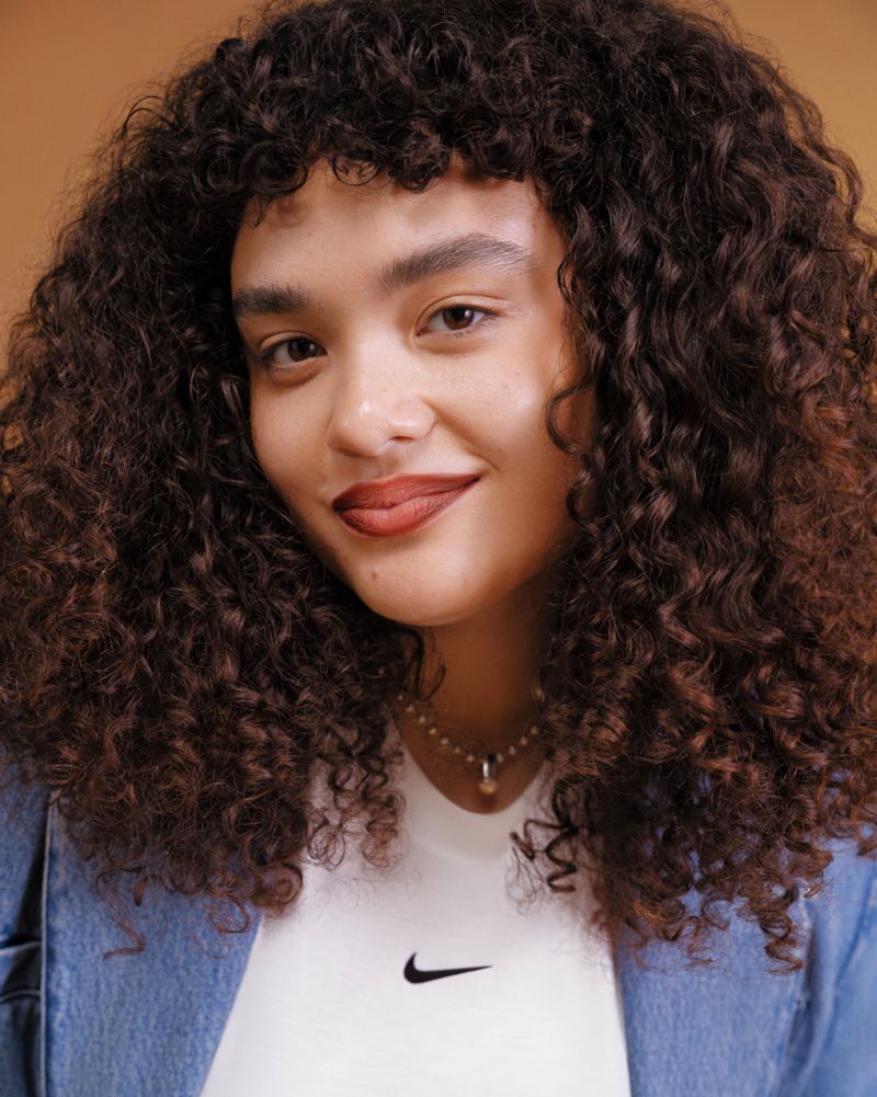 Portrait of a curly-haired young woman wearing a denim jacket and white Nike t-shirt against a neutral background, looking at the camera.