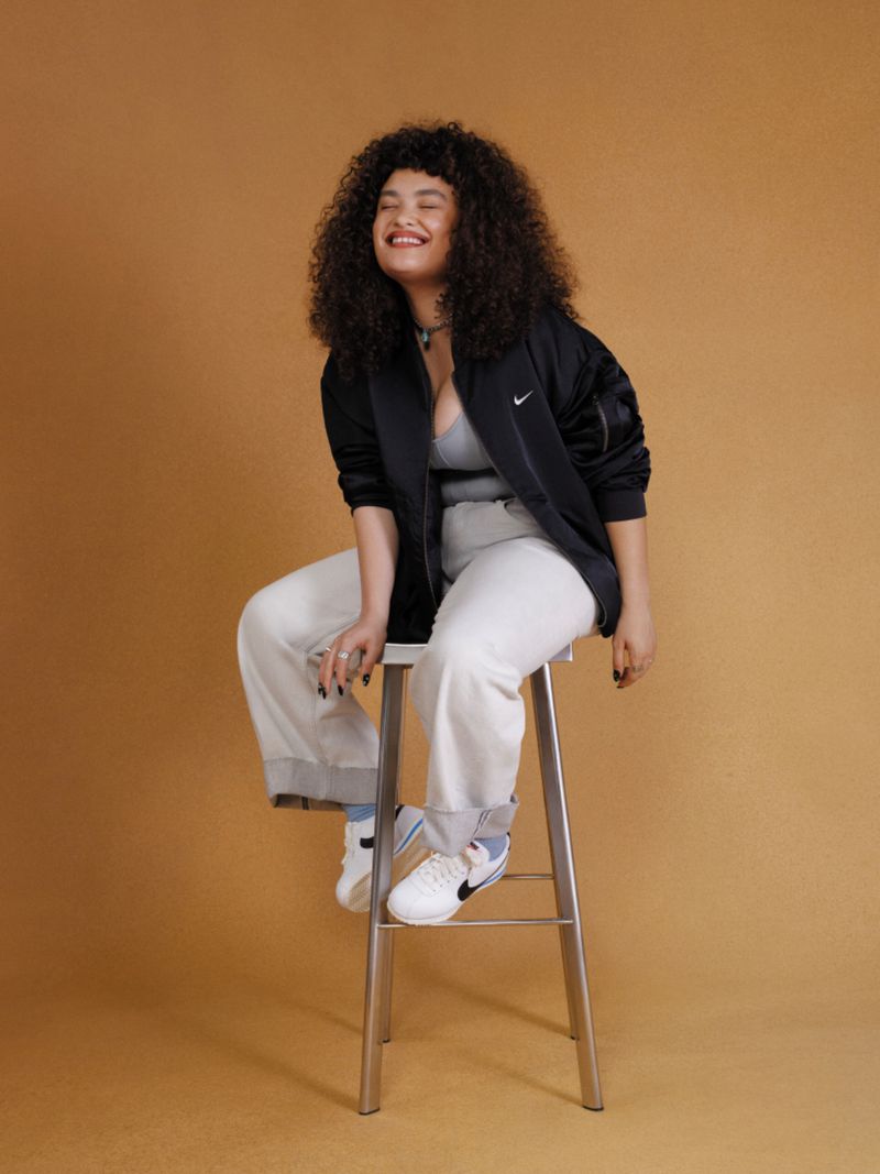 A curly-haired woman in a black jacket and white pants sits smiling on a stool in a studio, with a light brown background.