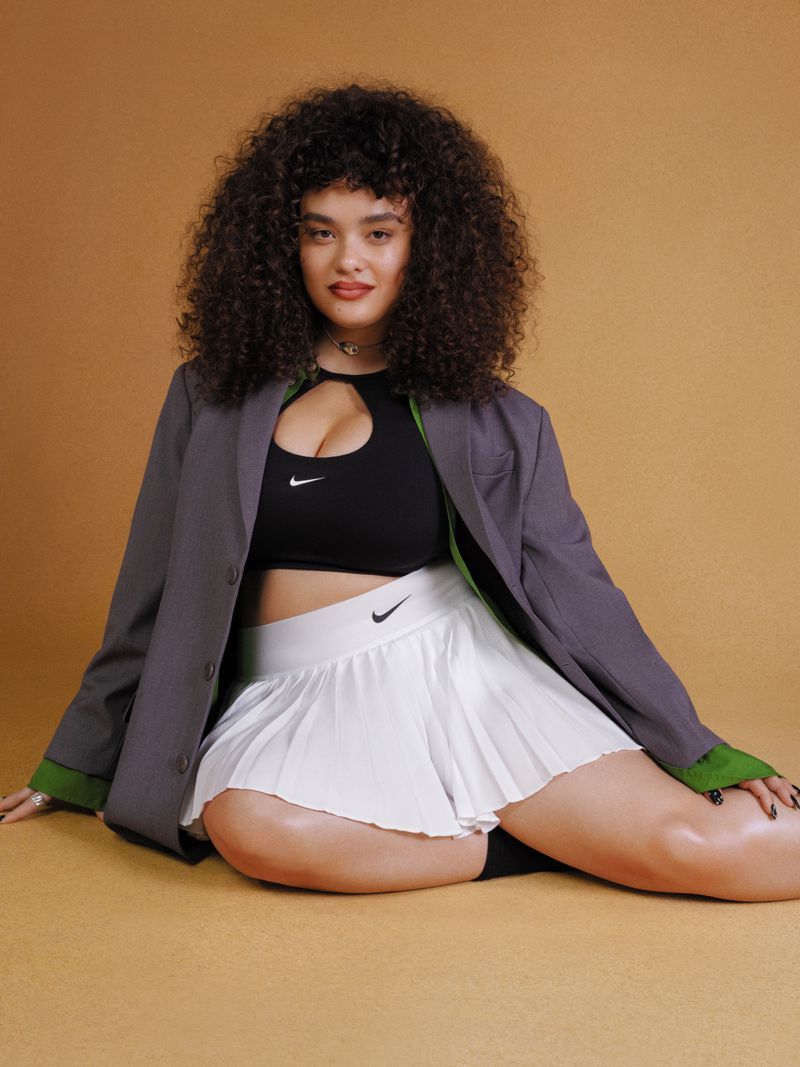 Woman with curly hair sits on a caramel surface wearing a black sports bra, white pleated skirt, and grey blazer in a photo studio.