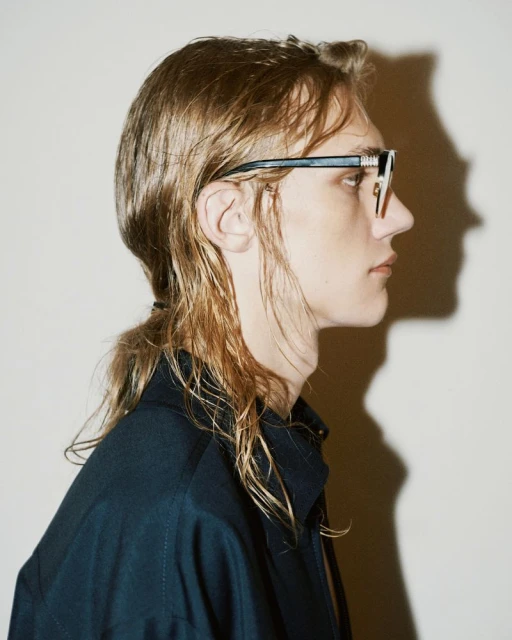 Profile portrait of a young adult man with blond, shoulder-length hair wearing glasses and a navy-blue collared shirt against a white background.