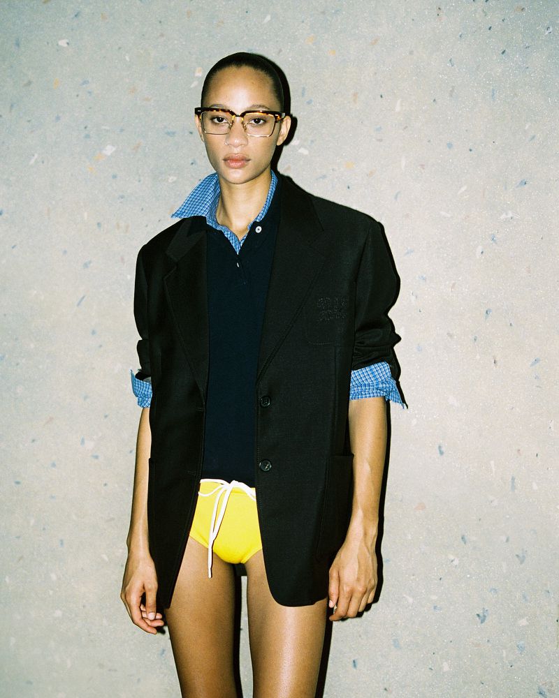 A woman with fair skin and glasses poses in a black blazer over a blue shirt, and yellow shorts against a speckled grey background.