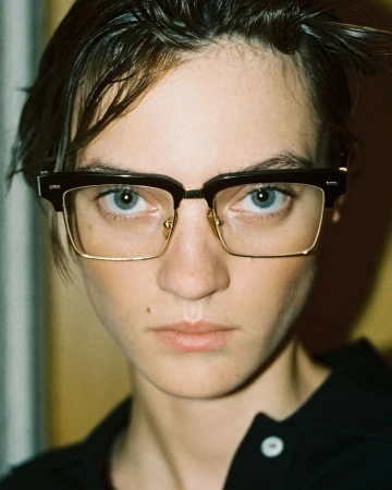 Close-up portrait of a model with light skin, wearing dark-framed glasses, looking directly at the camera, shot indoors with natural light.