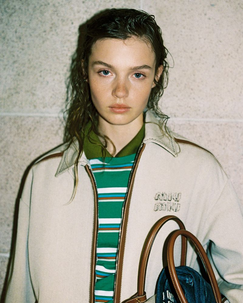 Close-up portrait of a young woman with wet dark hair wearing a collared jacket and striped shirt while carrying a tote bag.