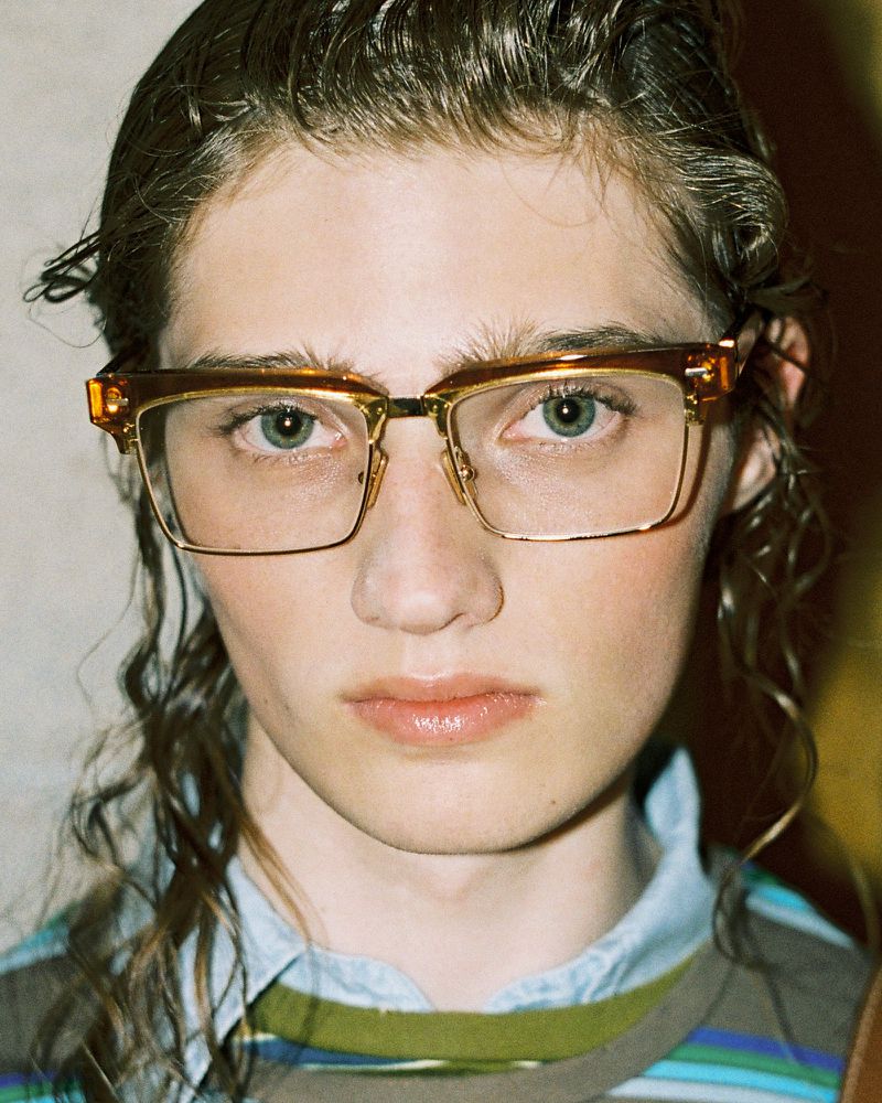 Close-up portrait of an adult wearing eyeglasses with a gold metal frame. They have light eyes, light skin, and wavy brown hair.
