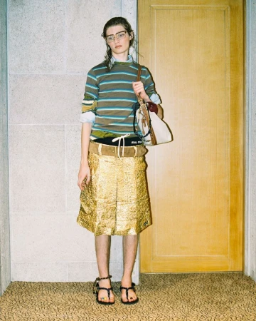 Woman in striped shirt and gold skirt stands in front of door, holding handbag. Fashion editorial shot with soft lighting and neutral setting.
