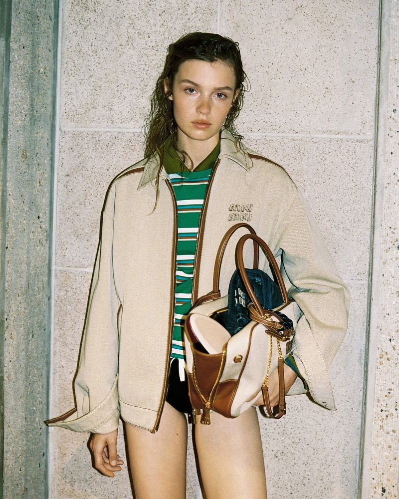 Model with damp hair wearing a light beige jacket over a green striped polo holding a structured handbag