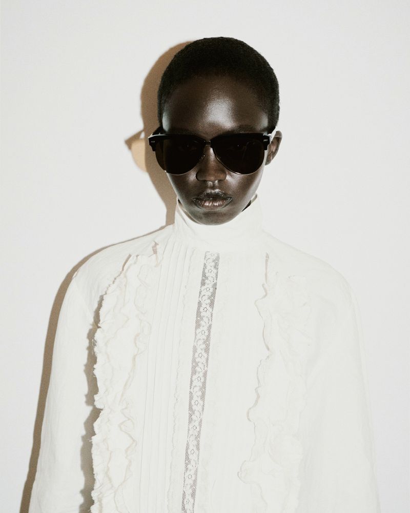 Model wearing dark sunglasses and a white ruffled high neck shirt against a plain light wall with a distinct shadow cast.