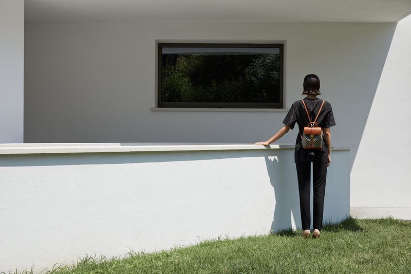 A woman stands on grass near a white building, facing away from the camera, wearing a backpack, on a bright sunny day.