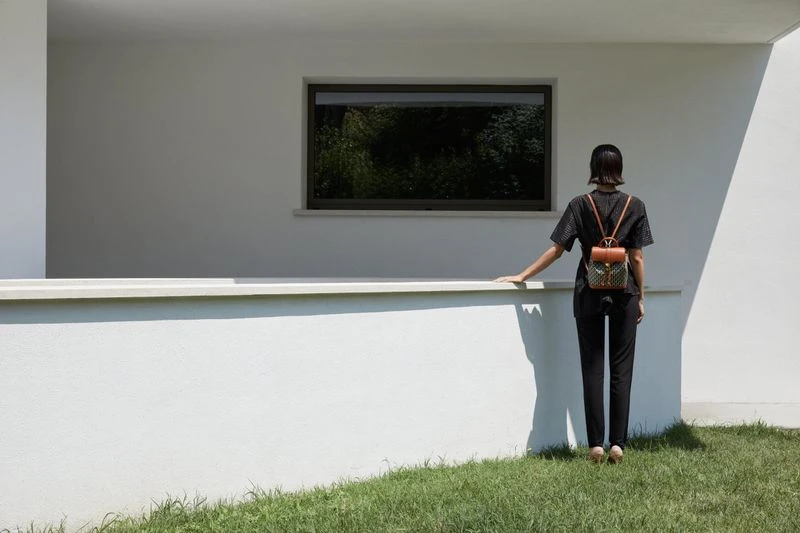 A woman stands on grass near a white building, facing away from the camera, wearing a backpack, on a bright sunny day.