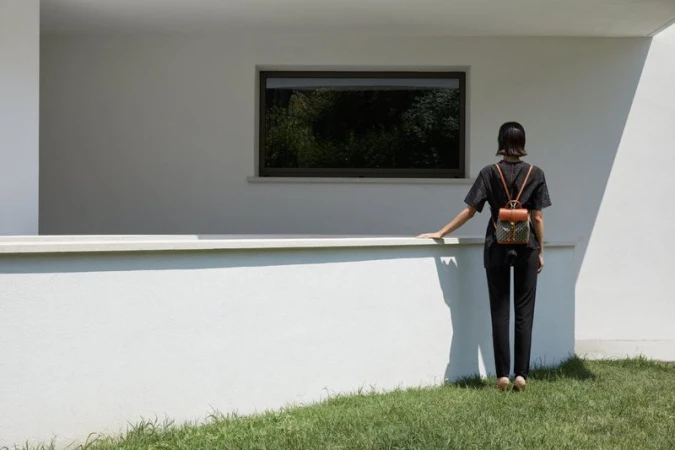 A woman stands on grass near a white building, facing away from the camera, wearing a backpack, on a bright sunny day.