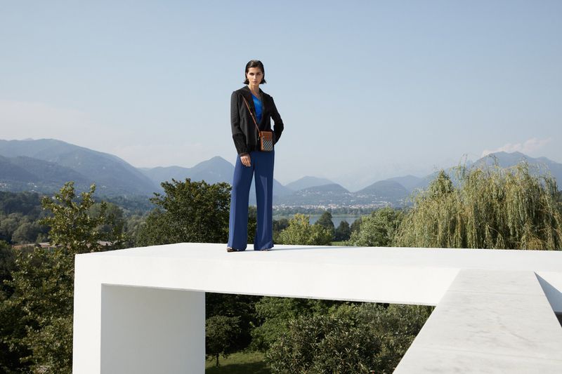 Adult woman in a jacket and blue pants poses outdoors on a white architectural structure against a mountain landscape backdrop.