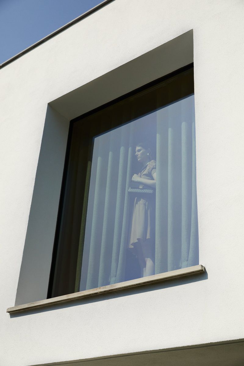 Window view of a woman in dress with arms crossed, standing behind vertical blinds in a white building on a sunny day.