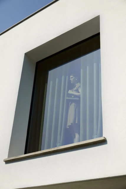 Window view of a woman in dress with arms crossed, standing behind vertical blinds in a white building on a sunny day.