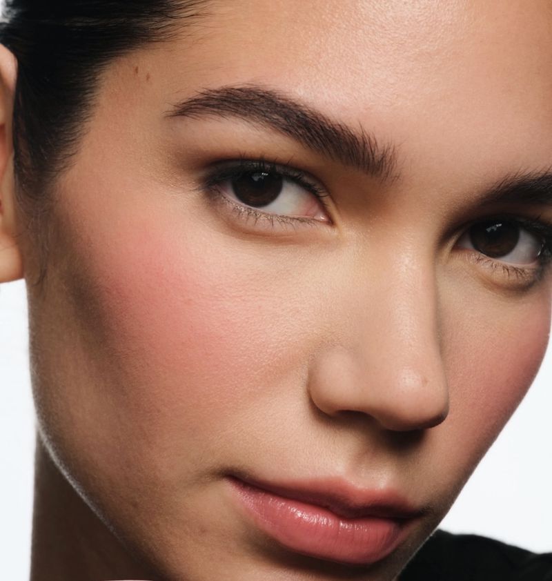 Close-up studio shot of a woman's face featuring makeup and skincare. Her skin has a pink blush. She has dark hair and eyebrows.