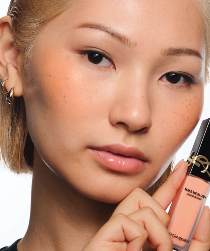 Close-up of an Asian woman with fair skin wearing blush and lip gloss, holding YSL makeup product against a white background.