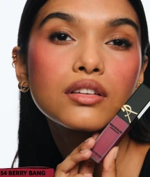 Close-up portrait of a model holding YSL Beauty Make Me Blush liquid blush against a white background