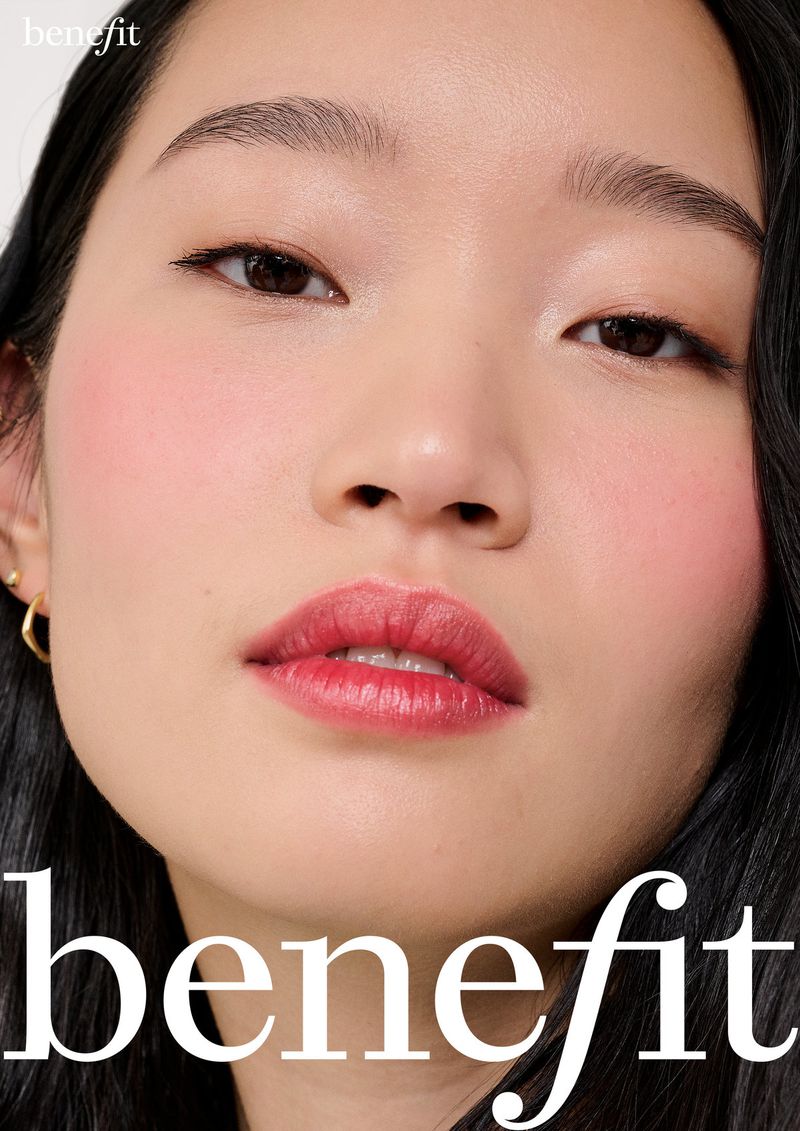 Close-up shot of an adult female model showcasing makeup, including blush and lipstick, in a beauty advertisement for Benefit Cosmetics.