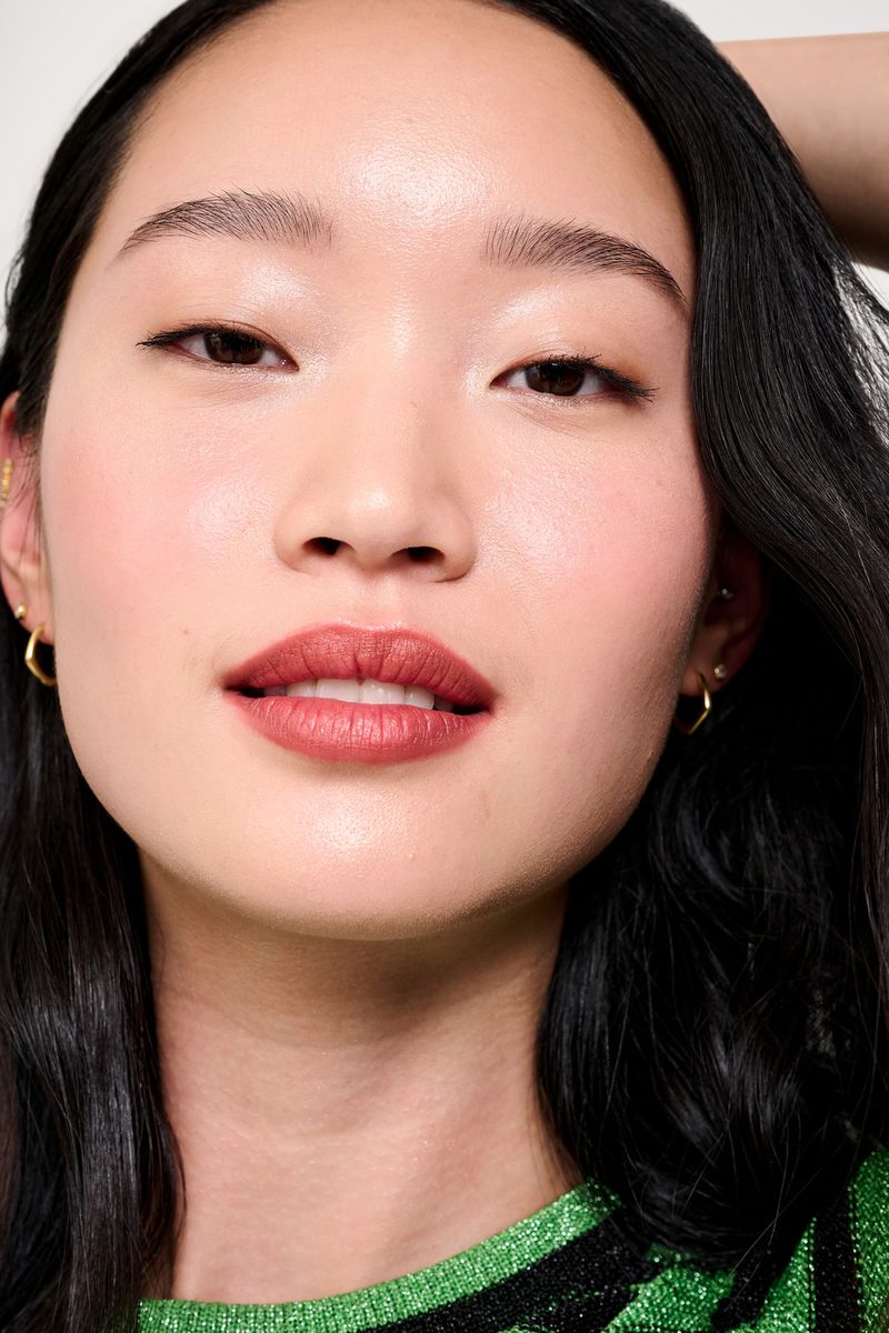 Close-up portrait of an Asian woman with dark hair, wearing a green shirt, and gold hoop earrings, against a bright white background.