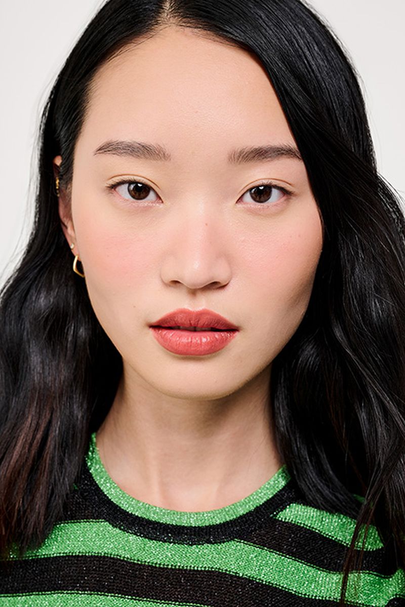 Close-up portrait of an Asian woman with dark hair and a green and black striped shirt, set against a plain white background.