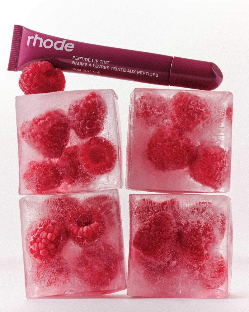 Close-up of skincare product (Rhode Peptide Lip Tint) on ice cubes containing raspberries, set against a clean white background.