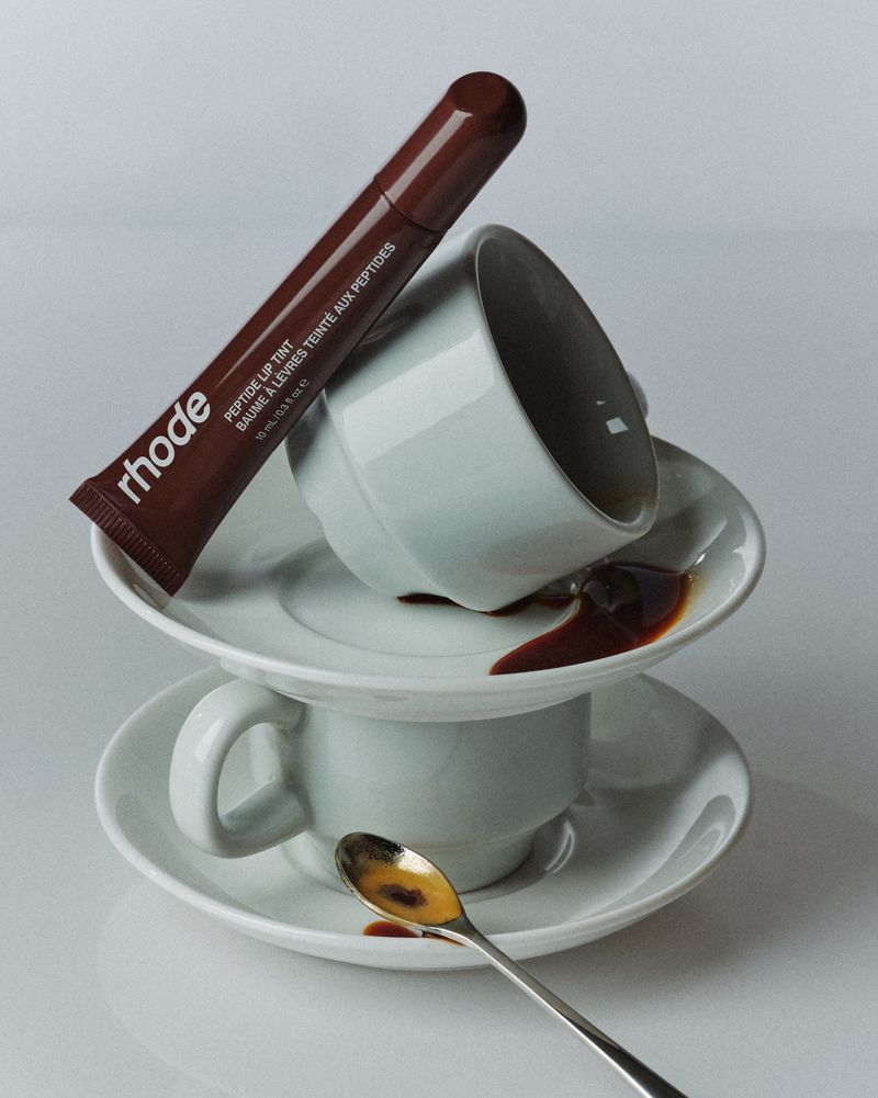 Still life of Rhode Peptide Lip Tint tube balanced on coffee cups and saucers, with spilled liquid on a white surface.