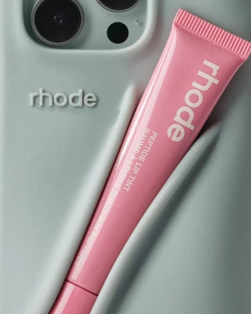 Close-up of a pink Rhode Peptide Lip Tint tube next to a light blue phone case with embossed 'rhode' logo and camera lens.