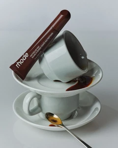 Brown Rhode Peptide Lip Tint tube balanced on stacked white ceramic espresso cups and saucers with spilled coffee.