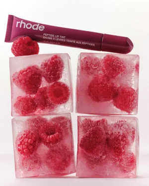 A dark berry tube of rhode Peptide Lip Tint rests on top of four large ice cubes containing frozen red raspberries against a white background.