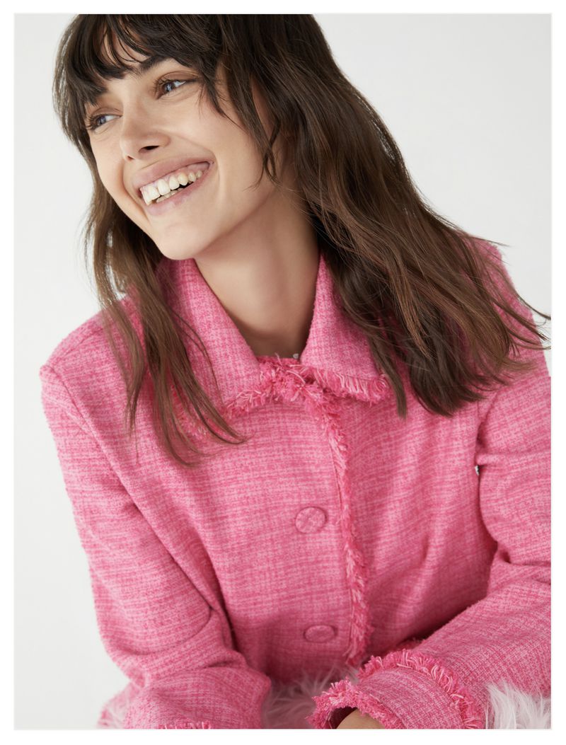 Brunette woman smiles wearing a pink tweed jacket with a collared neck, long sleeves, and button closure in a light background.